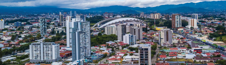 Aerial view of a bustling city with a mix of modern skyscrapers and residential buildings, featuring a large sports arena with a distinctive dome shape amidst the urban landscape, surrounded by lush greenery and distant mountains under a cloudy sky.