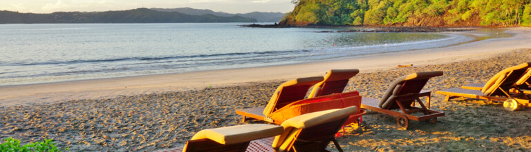 Several beach loungers are arranged on a sandy shore, facing a calm sea at sunset, with gentle waves lapping at the beach and lush greenery in the background.