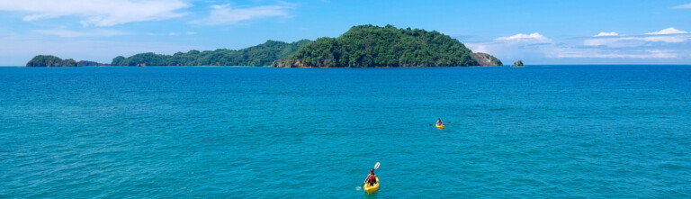 Two kayakers in yellow kayaks navigating through turquoise waters, with green islands in the background under a clear blue sky.