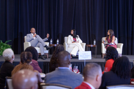 A panel discussion featuring three speakers on stage, with a man in a light gray suit speaking into a microphone, a woman in a white suit listening, and another woman in a maroon dress taking notes. The audience is mostly visible from the back, with several people seated at tables and a sign indicating 'Table 4 Black Alumni Council' in the foreground.