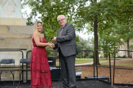 A woman in a burgundy dress is shaking hands with a gray-suited man while receiving an award on an outdoor stage surrounded by trees.
