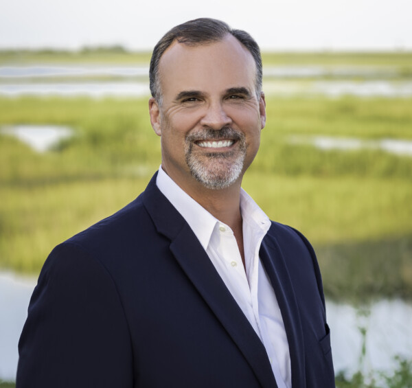 A smiling man in a dark blazer and white shirt stands outdoors with a background of lush green marshland and water.