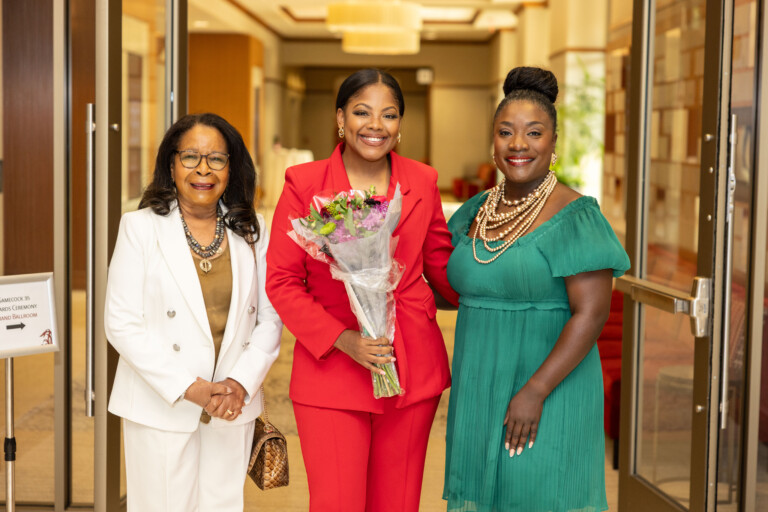 Three women stand together in a brightly lit entrance area, celebrating an occasion. The woman in the center wears a bright red suit and holds a bouquet of flowers, smiling widely. The woman on the left is dressed in white with a gold top and holds a small handbag, while the woman on the right wears a green dress and a statement necklace, also smiling. A sign for an awards ceremony is visible in the background.