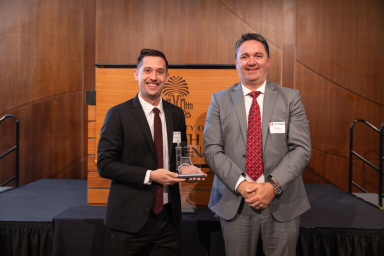 Two men are standing side by side, one holding an award, in a formal setting with wooden paneling and a podium in the background. The man on the left is dressed in a black suit with a white shirt and dark tie, while the man on the right is in a gray suit with a patterned tie. Both are smiling.