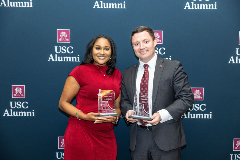 Two individuals stand smiling in front of a USC Alumni backdrop, holding awards. The person on the left wears a red dress and has long dark hair, while the person on the right is dressed in a suit and tie, displaying an award that reads 'GAMECOCK.' Both are celebrating an achievement.