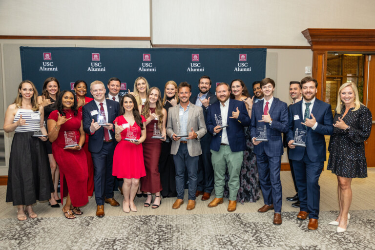 A group of diverse individuals poses together, holding awards at a USC Alumni event, with a backdrop featuring the USC Alumni logo. The attendees are dressed in formal attire, displaying excitement and camaraderie.