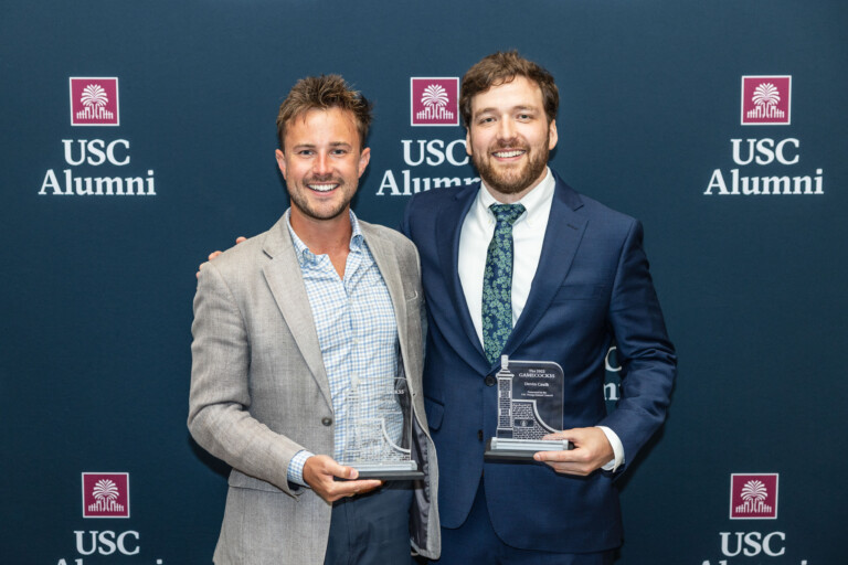 Two men stand together holding awards in front of a dark blue backdrop featuring the USC Alumni logo. The man on the left wears a light gray blazer over a checkered shirt, while the man on the right is dressed in a navy suit with a patterned tie.