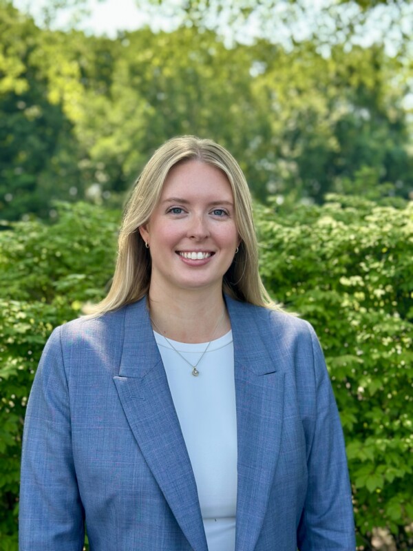 A smiling woman with long blonde hair, wearing a light blue blazer over a white top, stands in front of lush green foliage on a sunny day.