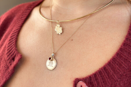 Close-up of a woman's neck featuring two gold necklaces: a thick choker and a layered pendant necklace, which highlights a round pendant with a red gemstone and an intricate leaf-shaped charm, all set against a background of a burgundy sweater.