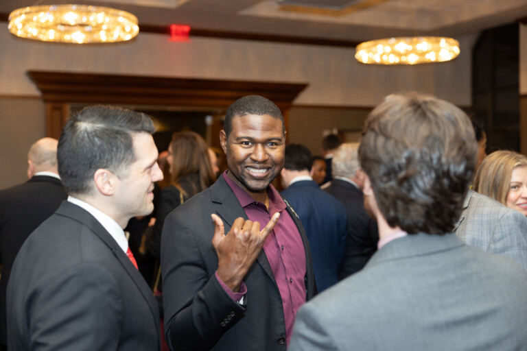 A man with a friendly smile, wearing a black blazer over a maroon shirt, is making a hand gesture in a social gathering filled with people in formal attire. The background features blurred figures and warm lighting from chandeliers.