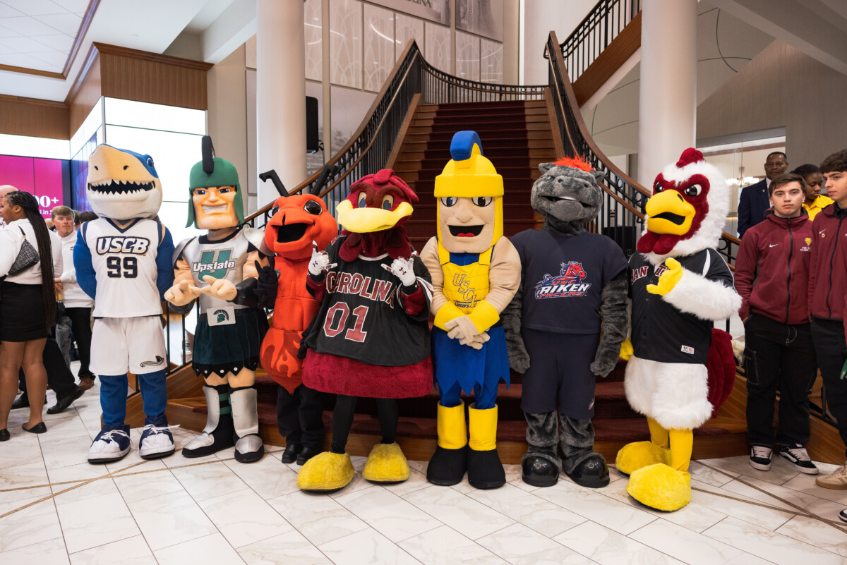 A group of seven colorful mascots stand on a staircase in an indoor setting, showcasing various collegiate themes with distinct costumes such as a shark, a Spartan warrior, a red bird, and a dragon. Some mascots are also dressed in sports jerseys. Nearby, a few individuals in casual attire observe the scene.