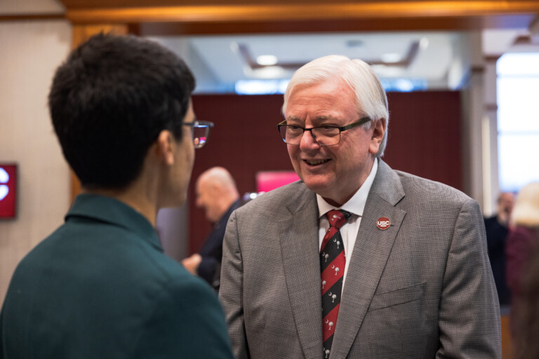 A man with gray hair and glasses, wearing a suit and a tie with a red and black pattern, smiles while engaged in conversation with another person, who is mostly out of frame and wearing a dark green jacket. The background features a softly blurred indoor setting with people mingling.