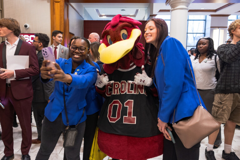 Two women in blue blazers pose for a selfie with a mascot in a black jersey reading 'CAROLINA 01', as a crowd of people in formal attire gather in the background of a bright indoor setting.