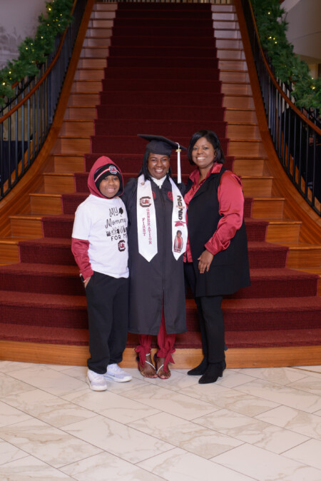 Three individuals standing on a staircase: a graduate in a cap and gown holding a graduation sash, flanked by two women—one in a white shirt with a graphic and red hoodie, and the other in a formal red blouse.