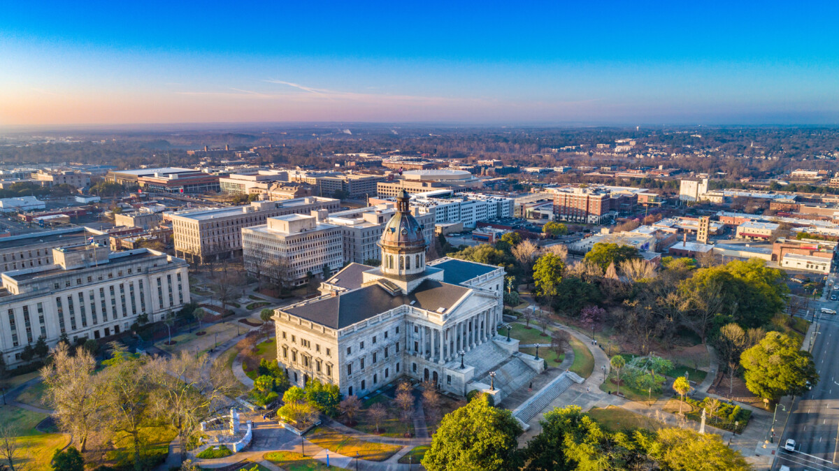 Aerial view of a historic government building with a dome, surrounded by green parks and downtown buildings in the background under a clear blue sky.