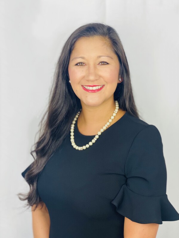 A smiling woman with long dark hair wearing a black dress with ruffled sleeves and a pearl necklace, set against a white background.