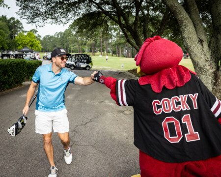 A man in a blue polo and white shorts shakes hands with a large red mascot named Cocky, set on a golf course.