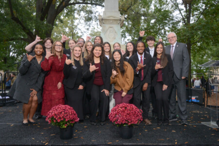 A large group of smiling people, dressed in professional attire, pose together with a white stone monument in the background, surrounded by flowering plants.
