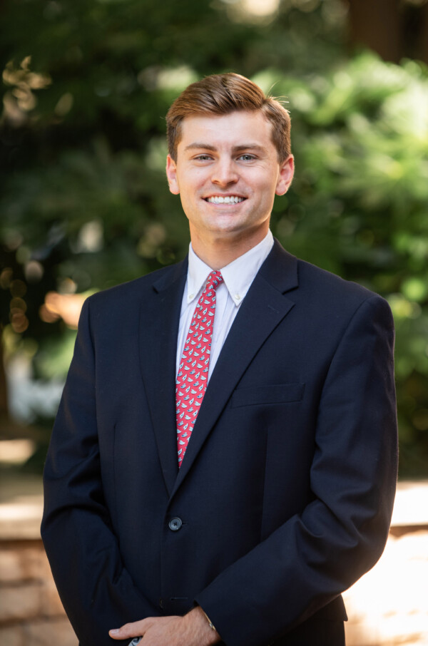 A young man in a dark suit and a patterned red tie smiles, standing outdoors with greenery in the background.