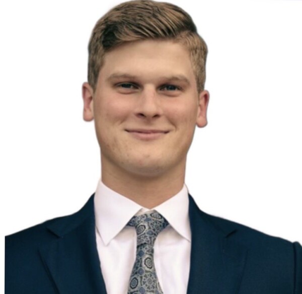 A young man with short blonde hair, wearing a dark suit and a patterned tie, smiling against a white background.