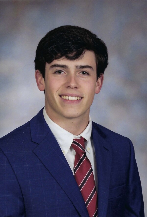 Young man with dark hair wearing a blue suit and a striped tie, smiling against a soft, neutral background.
