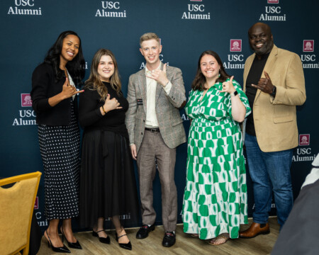 A group of five USC alumni poses together, smiling, with a backdrop featuring the USC Alumni logo.
