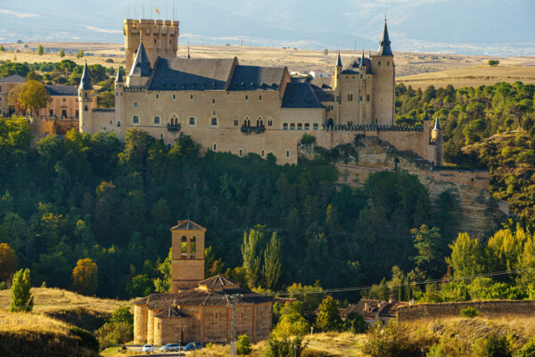 A medieval castle with prominent towers and battlements sits atop a hill, surrounded by lush greenery and distant mountains.