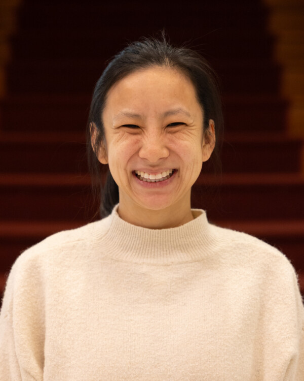A woman with long dark hair, wearing a cream-colored sweater, smiling warmly against a blurred background of stairs.
