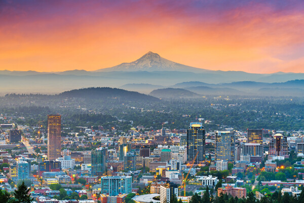 A panoramic view of a city skyline with Mount Hood in the background, set against a colorful sunset sky.