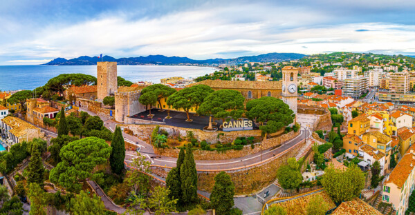 Aerial view of Cannes featuring a historical tower, lush trees, and the coastline with hills in the background.