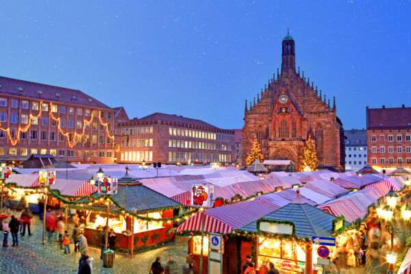 A festive Christmas market filled with colorful stalls, twinkling lights, and a large church in the background, under a snowy sky.