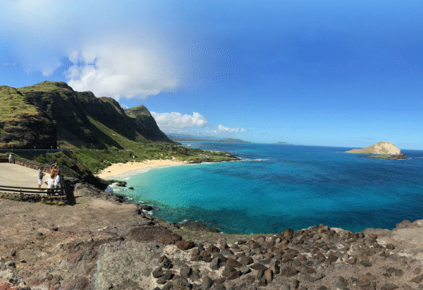 A panoramic view of a coastal landscape featuring green mountains, a sandy beach, and turquoise water under a bright blue sky.