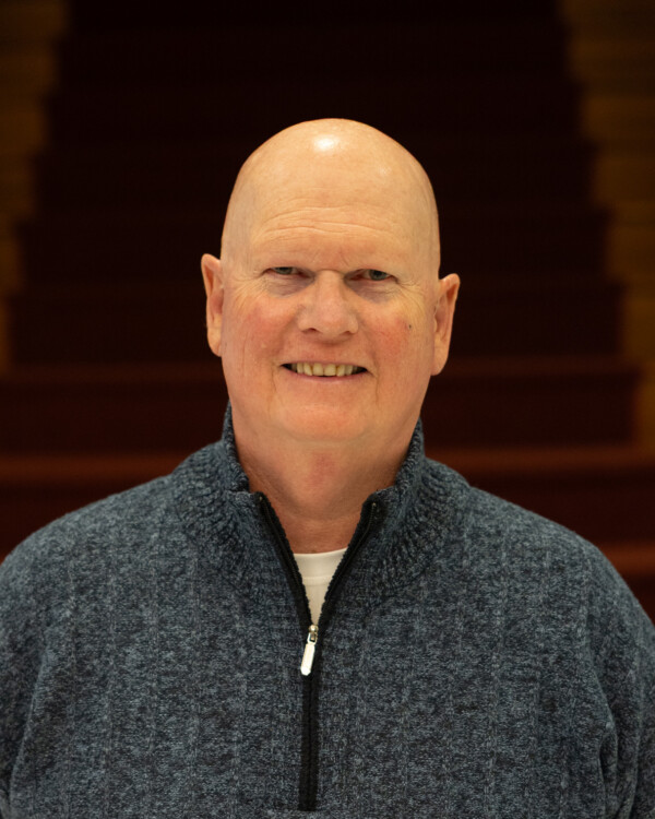 A smiling man with a bald head wearing a blue sweater, standing in front of a wooden staircase.