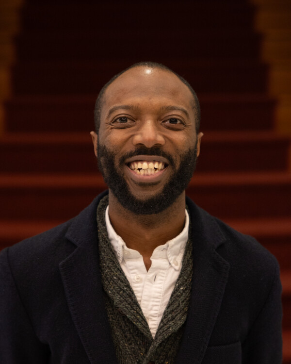 Smiling man with a beard and short hair, wearing a white shirt and a dark blazer, stands in front of red stairs.