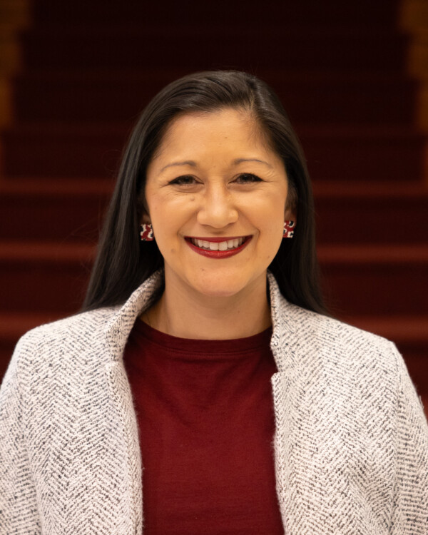 A woman with long black hair and a bright smile, wearing a maroon shirt and a light-colored cardigan, stands in front of wooden stairs.