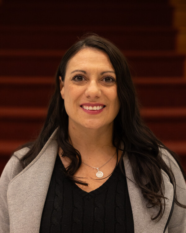 A smiling woman with long dark hair is wearing a black sweater and a light gray coat, standing against a backdrop of red stairs.