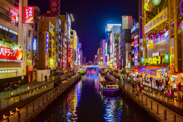 Vibrant cityscape at night featuring colorful neon signs, bustling streets, and a canal with boats along the waterway.