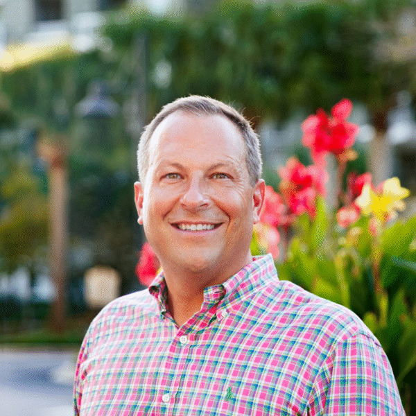 Smiling man in a checkered shirt stands outdoors, surrounded by vibrant flowers and greenery.