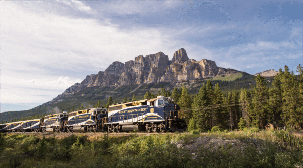 A Rocky Mountaineer train travels alongside a lush forest, with rugged mountain peaks towering in the background under a blue sky.