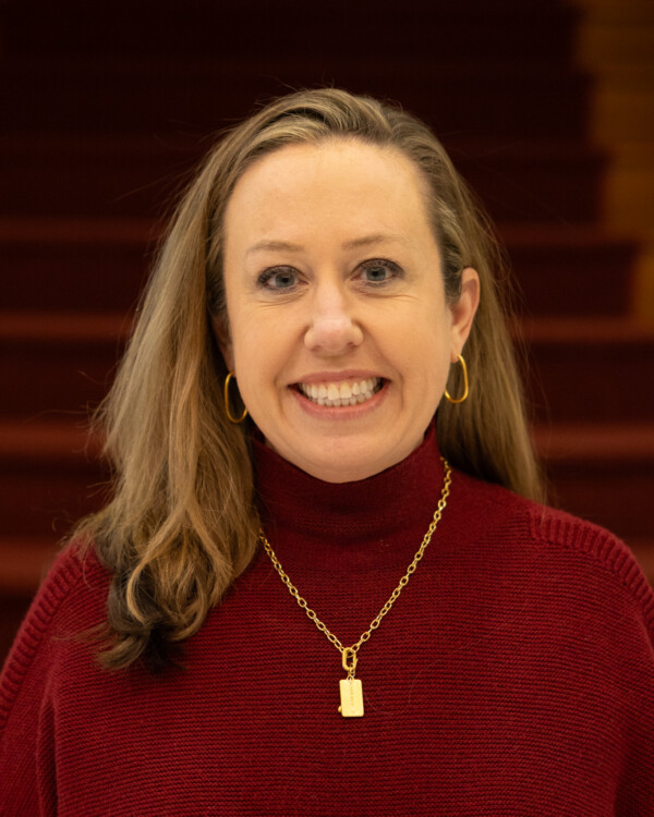 A woman with long brown hair wearing a burgundy turtleneck and gold jewelry, smiling in a setting with wooden stairs behind her.