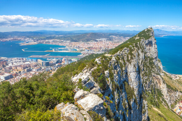A panoramic view of the Rock of Gibraltar overlooking the city and harbor, with lush greenery at the peak and a clear blue sky.