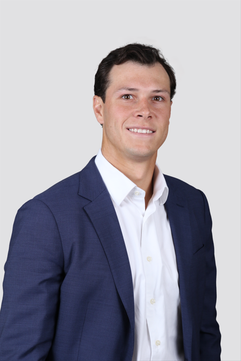 A young man in a dark blue suit and white shirt smiles confidently against a plain gray background.