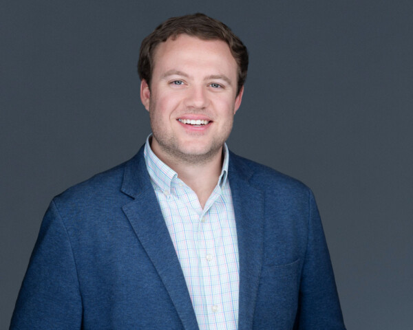 A man smiling, wearing a blue blazer over a checkered shirt, stands against a gray background.