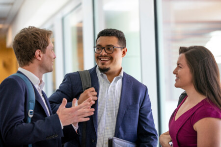 Three professionals engaged in conversation in a modern office setting, smiling and dressed in business attire.