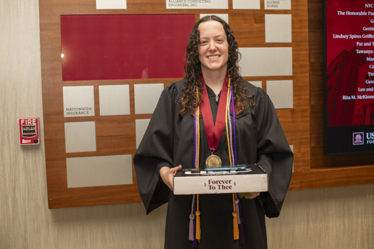 A graduate in a black cap and gown holds a tray with a certificate and pens, standing in front of a display board.