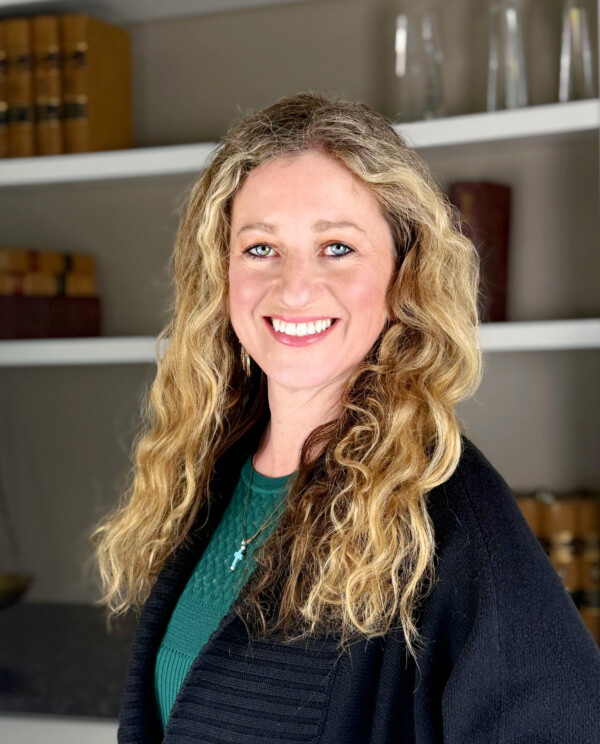 A woman with long, wavy blonde hair smiles warmly, wearing a black sweater over a green top, in a well-lit indoor setting.