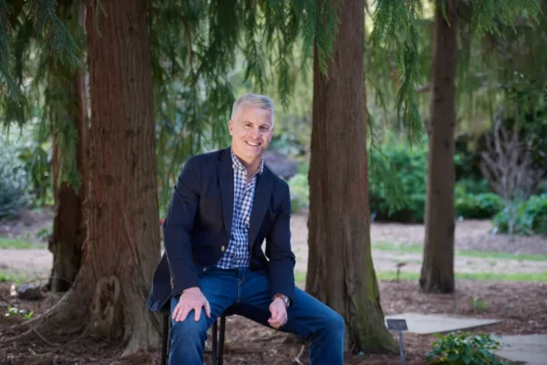 A smiling man with short gray hair sits on a stool among tall trees in a green garden setting, wearing a blazer and checkered shirt.