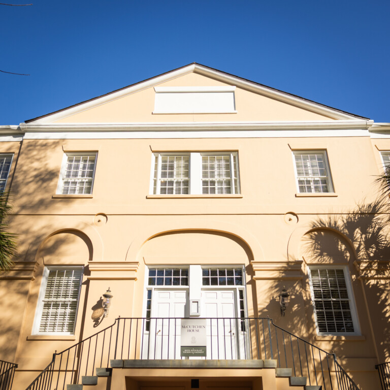 A pale yellow building with white windows and doors, featuring a staircase and palm trees nearby under a clear blue sky.