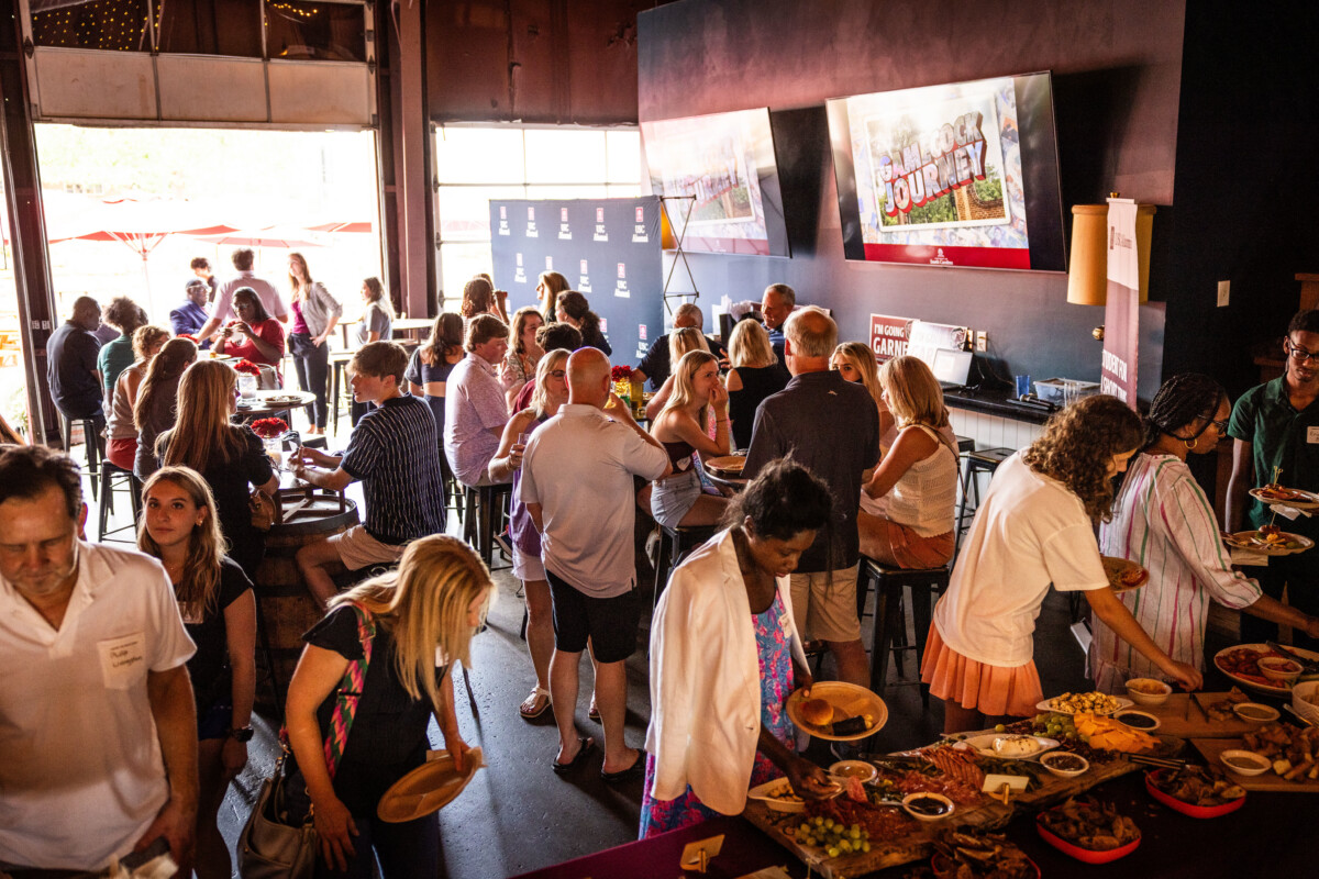 A crowded event inside a venue, with people mingling, enjoying food, and interacting at tables, surrounded by large screens.
