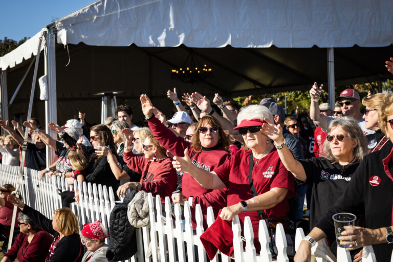 A crowd of enthusiastic fans, dressed in red and black, raises their arms in celebration at an outdoor event, surrounded by a white picket fence.